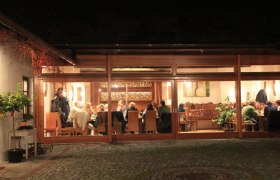 Heurigenlokal, © Ecker Ein gemütliches Heurigenlokal mit Gästen, die an Tischen sitzen, durch große Fenster sichtbar.