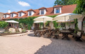 Weingut Burger, © Fam. Burger Ein gemütlicher Innenhof eines Weinguts mit Holztischen, Bänken und Sonnenschirmen, umgeben von einem Gebäude mit roten Ziegeldächern und grüner Bewachsung.