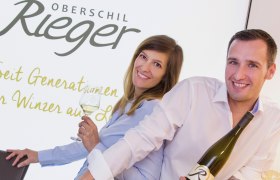 Heuriger Oberschil-Rieger, © Oberschil-Rieger Zwei Personen lächeln und halten Weinflaschen vor einem Schild mit der Aufschrift 'Oberschil Rieger'.