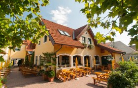 Schimmelbauer, © Baumgartner J. Ein gelbes Gebäude mit roten Dachziegeln und einem Biergarten mit Holzbänken und Sonnenschirmen.