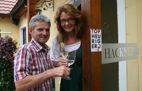 Winzerhof Hackner, © Hackner Ein Mann und eine Frau stehen vor einem Gebäude mit einem Schild 'Top Heuriger' und halten Weingläser.
