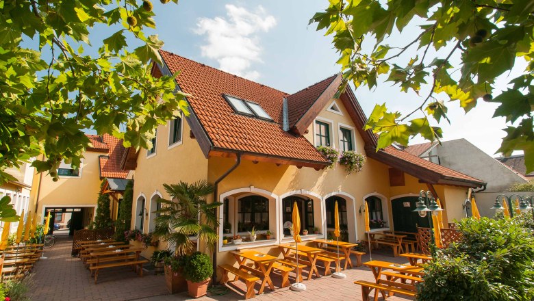 Schimmelbauer, © Baumgartner J. Ein gelbes Gebäude mit roten Dachziegeln und einem Biergarten mit Holzbänken und Sonnenschirmen.