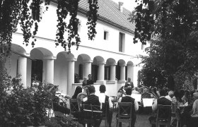 Veranstaltung beim Heurigenbetrieb, © Fam Dietzl Eine Gruppe von Menschen sitzt im Freien vor einem Gebäude mit Säulen, während eine Blaskapelle spielt.