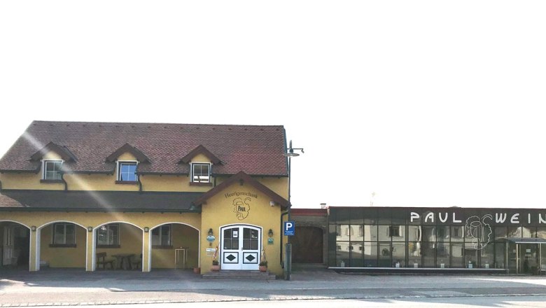 Heurigenlokal Paul, © Weingut-Heuriger Paul Außenansicht eines gelben Heurigenlokals mit dem Namen 'Paul' und einem modernen Anbau mit der Aufschrift 'Paul Wein'.