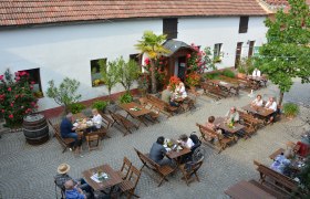 Heuriger im Innenhof, © Sophie Kienberger Menschen sitzen an Holztischen in einem gepflasterten Innenhof eines Heurigen, umgeben von Blumen und Bäumen.