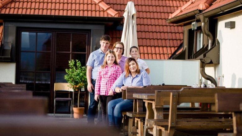 Familie Kropf, © Alexander Pfeffel Eine Familie posiert auf einer Terrasse mit Holztischen und roten Dachziegeln im Hintergrund.