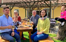 Familie Baumgartner, © Der Baumgartner Eine Familie sitzt an einem Holztisch im Freien und genießt eine Mahlzeit. Ein Kind sitzt auf einem Baumstamm. Im Hintergrund ein gelbes Haus mit Pergola.