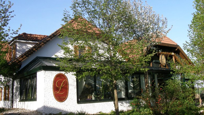 Kellertriftheuriger Loiskandl Aussenansicht, © Pfeifer Außenansicht eines traditionellen Gebäudes mit roten Dachziegeln und einem großen L-Logo an der Wand, umgeben von Bäumen und Pflanzen.