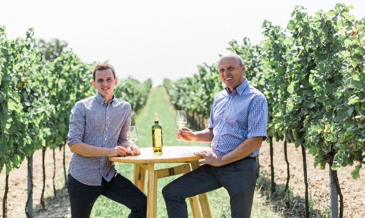 Familie Paul, © Weingut-Heuriger Paul Zwei Männer sitzen an einem Tisch im Weinberg mit Weingläsern.