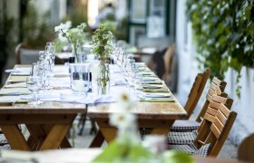 Gebeshuber, © Gebeshuber Langer Holztisch im Freien, gedeckt mit Gläsern, Besteck und Blumen.