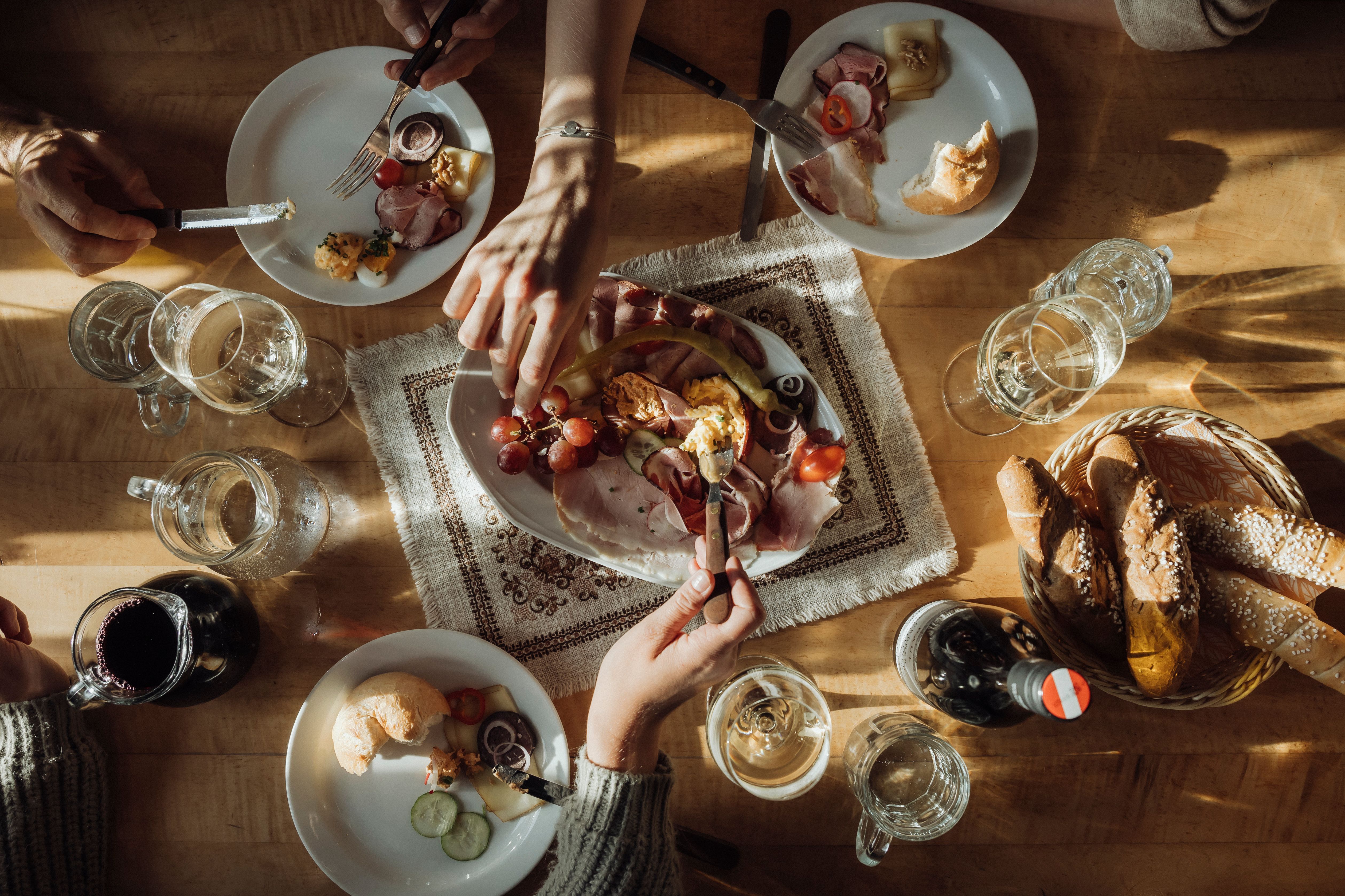 Ein Tisch mit einer Heurigenjause, bestehend aus Brot, Aufschnitt, Käse und Wein, umgeben von mehreren Händen.