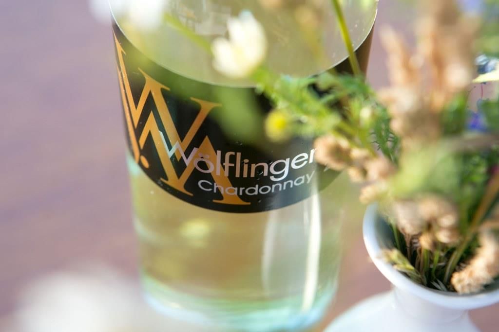 Nahaufnahme einer Weinflasche mit der Aufschrift 'Wölflinger Chardonnay' neben einem Blumenstrauß.