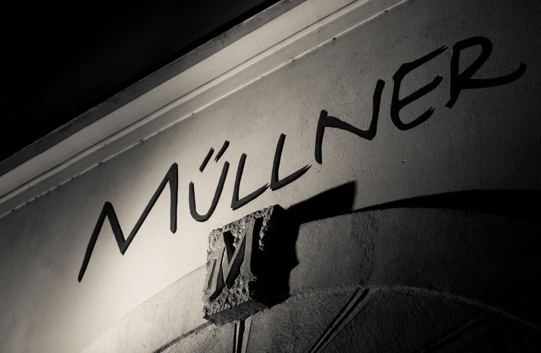 Schwarz-weißes Foto eines Schriftzugs 'Müllner' auf einer Wand mit einem dekorativen 'M'.