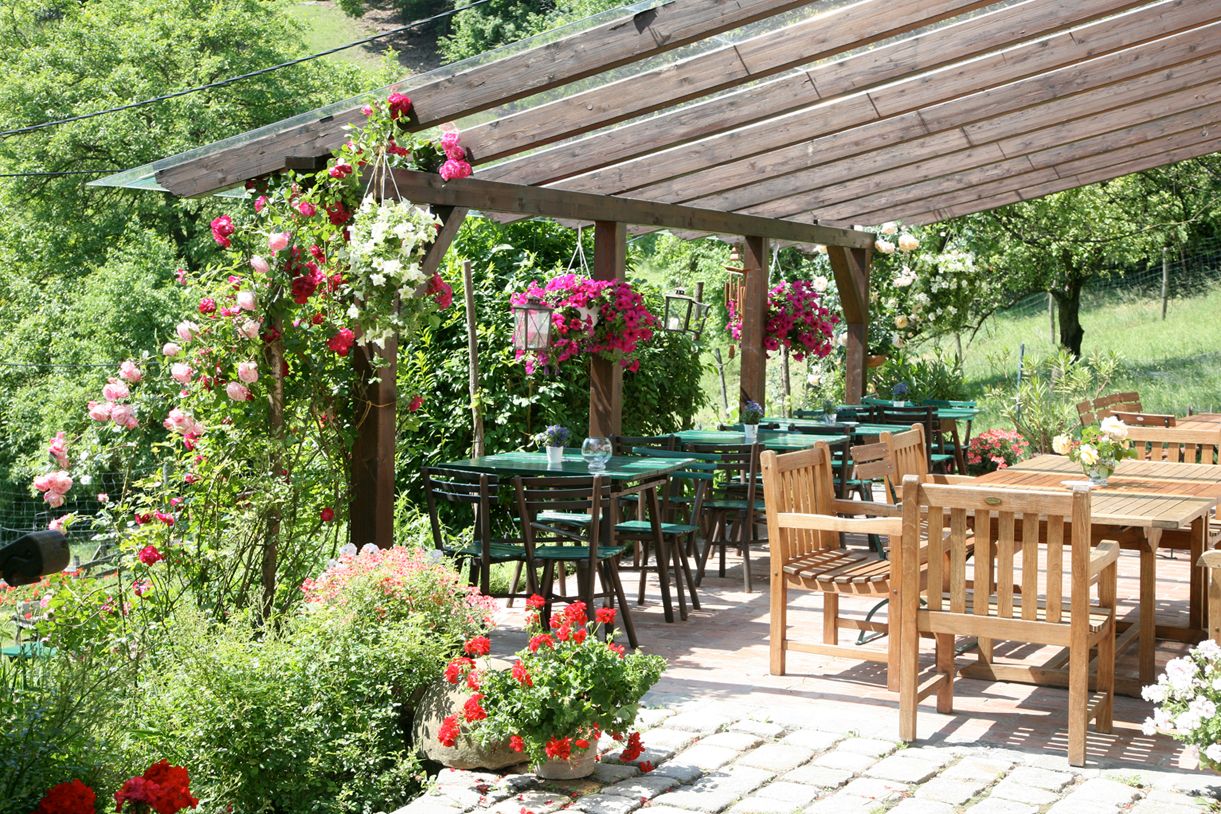 Ein gemütlicher Außenbereich eines Heurigen mit Holztischen und -stühlen, umgeben von blühenden Pflanzen und einem hölzernen Pergola-Dach.