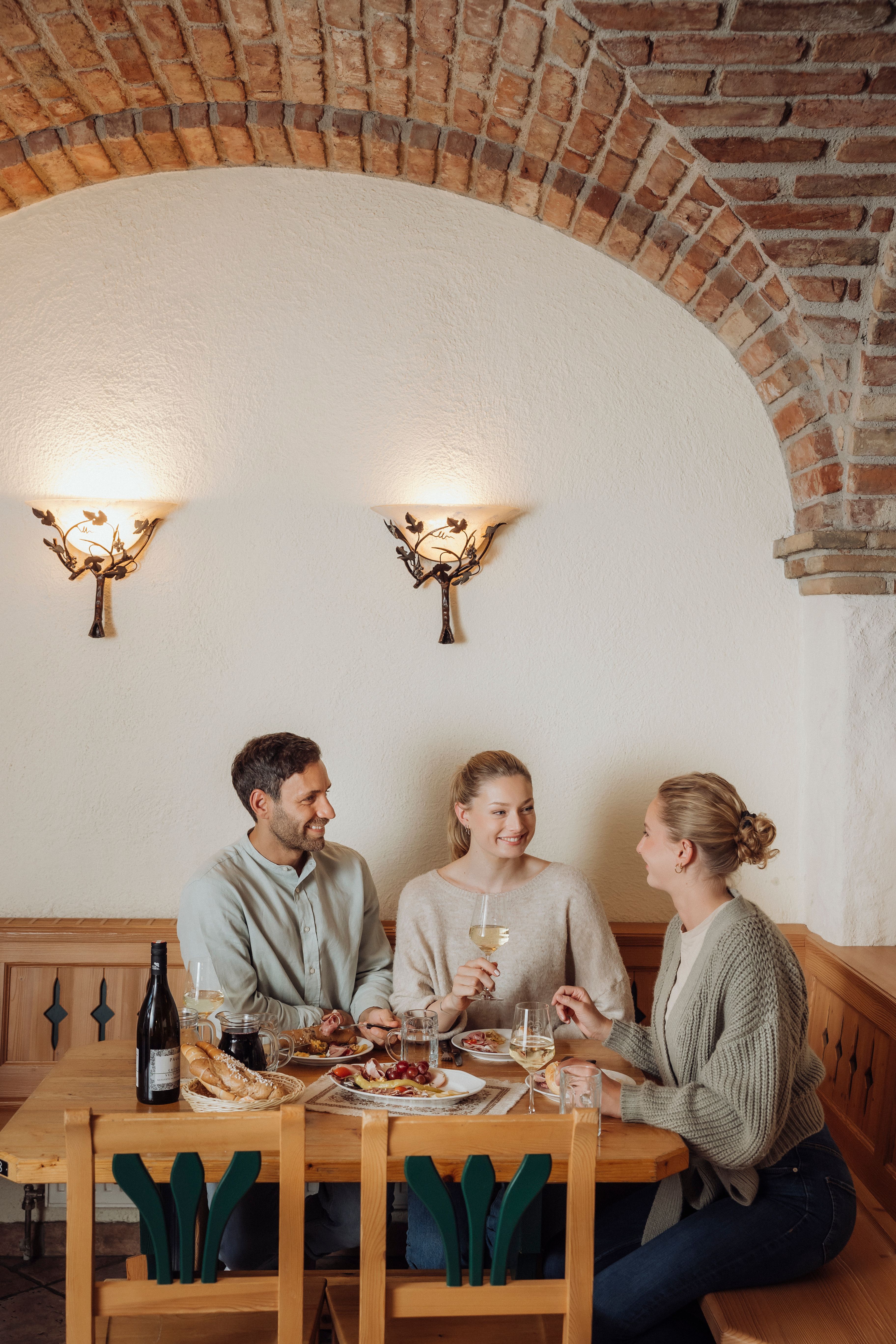 Drei Personen sitzen an einem Holztisch in einem Weinkeller und genießen Wein und Essen.
