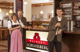 Drei Personen stehen an einer Bar im Weingut Schlossberg mit Weingl&auml;sern in der Hand.