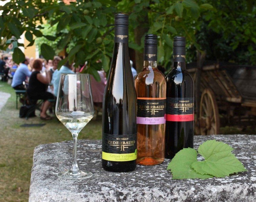 Drei Weinflaschen und ein Glas auf einem Tisch im Freien, mit Menschen im Hintergrund.