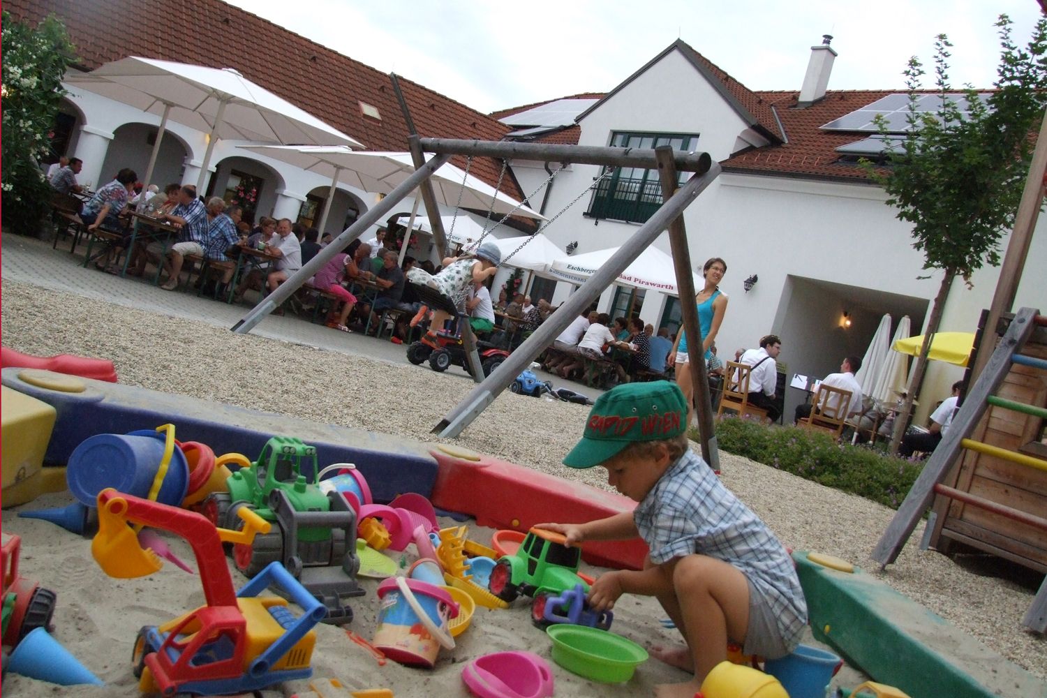 Ein Kind spielt in einem Sandkasten mit buntem Spielzeug. Im Hintergrund sitzen Menschen an Tischen unter Sonnenschirmen.