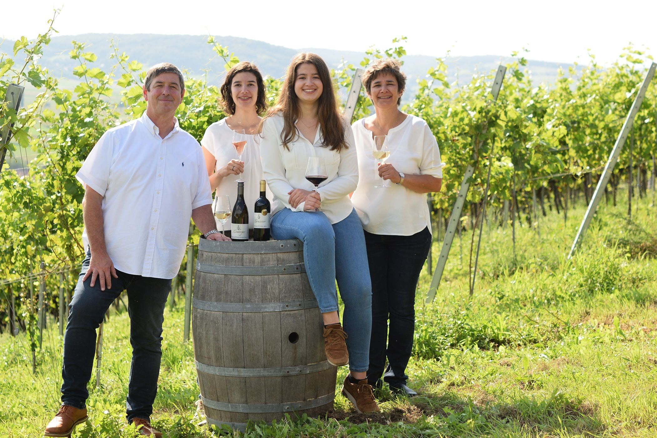 Vier Personen stehen und sitzen in einem Weinberg, lächelnd mit Weingläsern in der Hand, neben einem Weinfass mit Flaschen darauf.