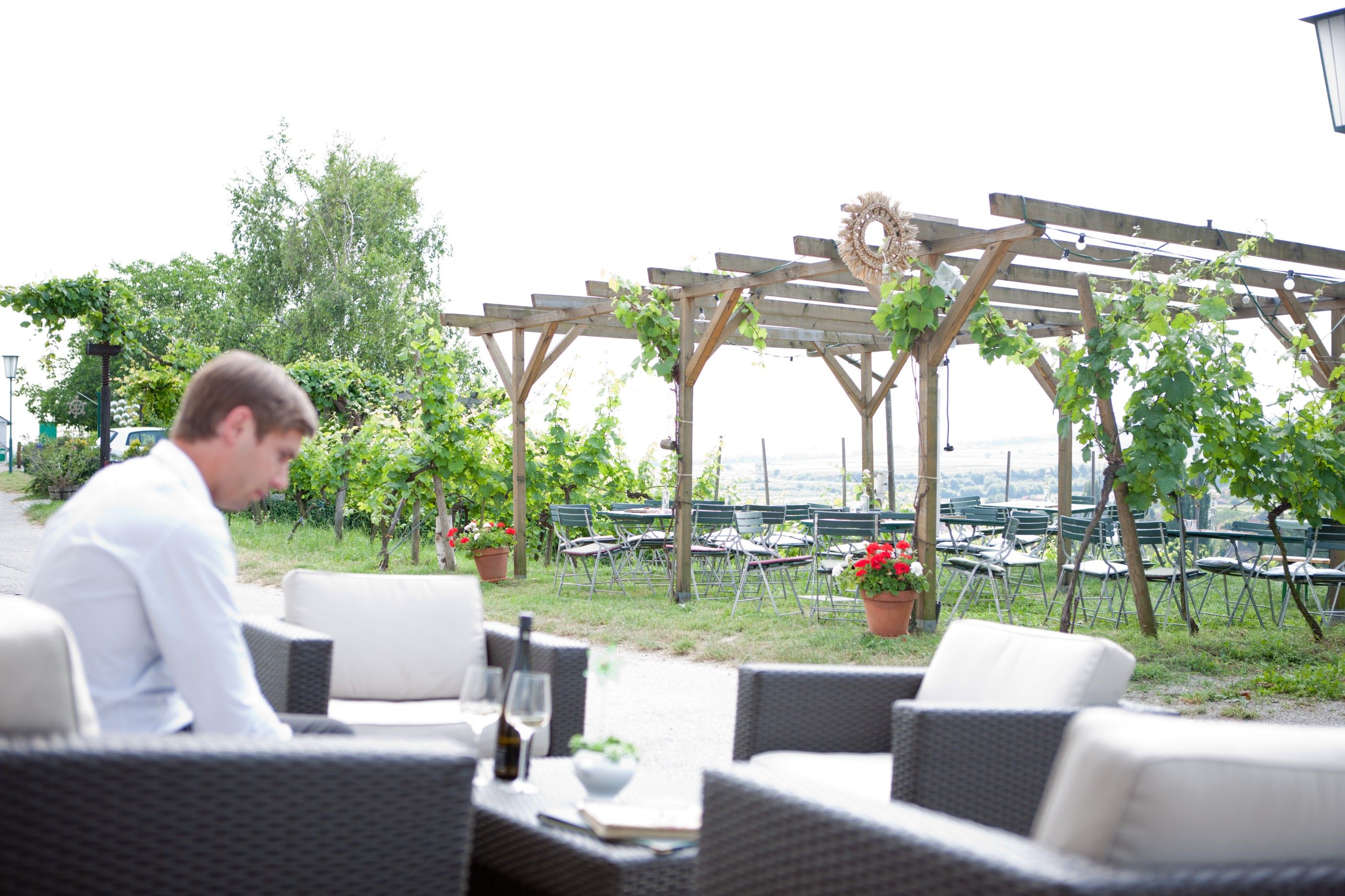 Ein Mann sitzt in einem Gastgarten mit Weinreben und leeren Tischen im Hintergrund.