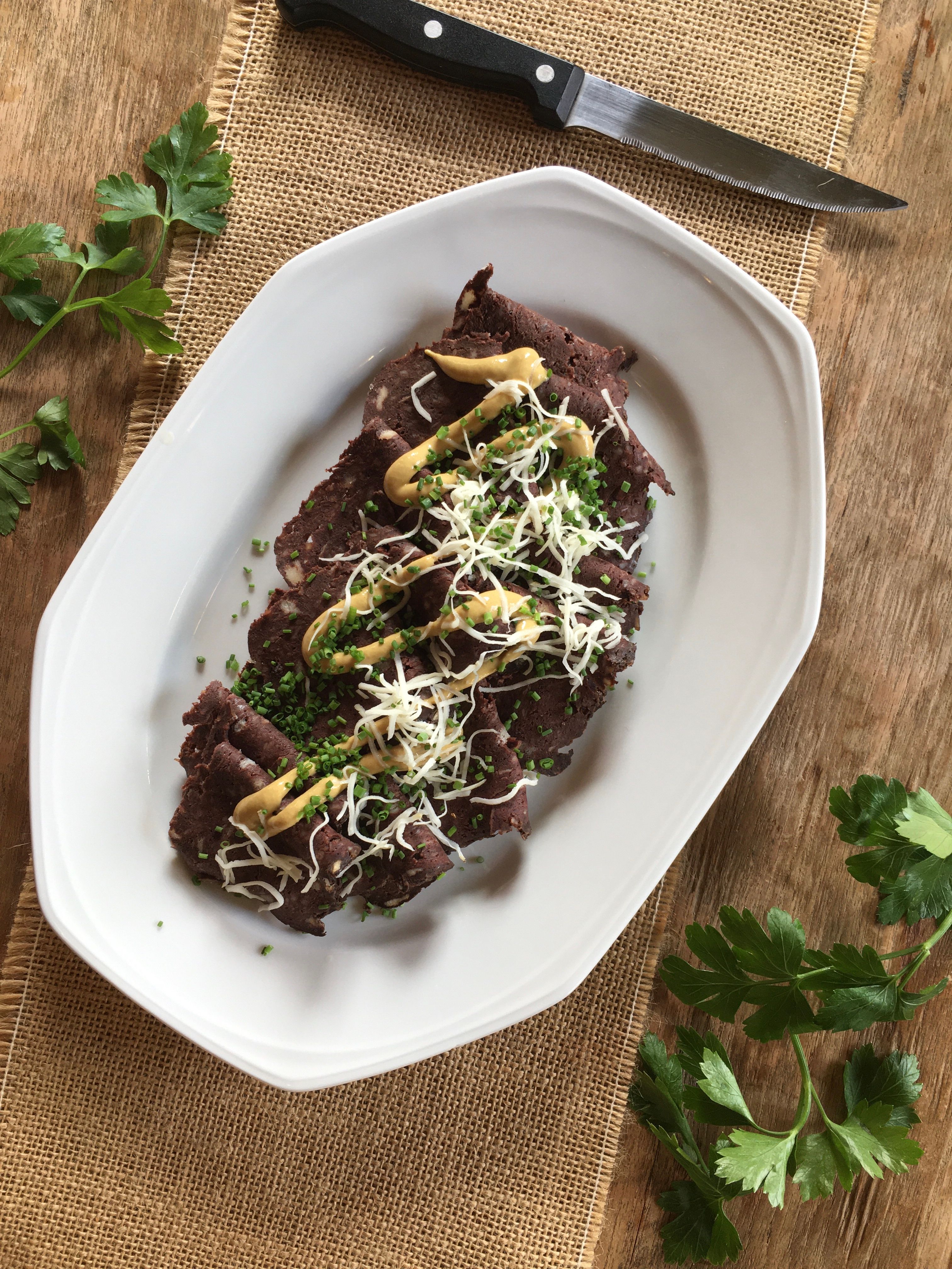 Ein Teller mit Blunzenbrot, garniert mit Senf, Schnittlauch und geriebenem Käse, auf einem Holztisch mit Petersilie und einem Messer daneben.