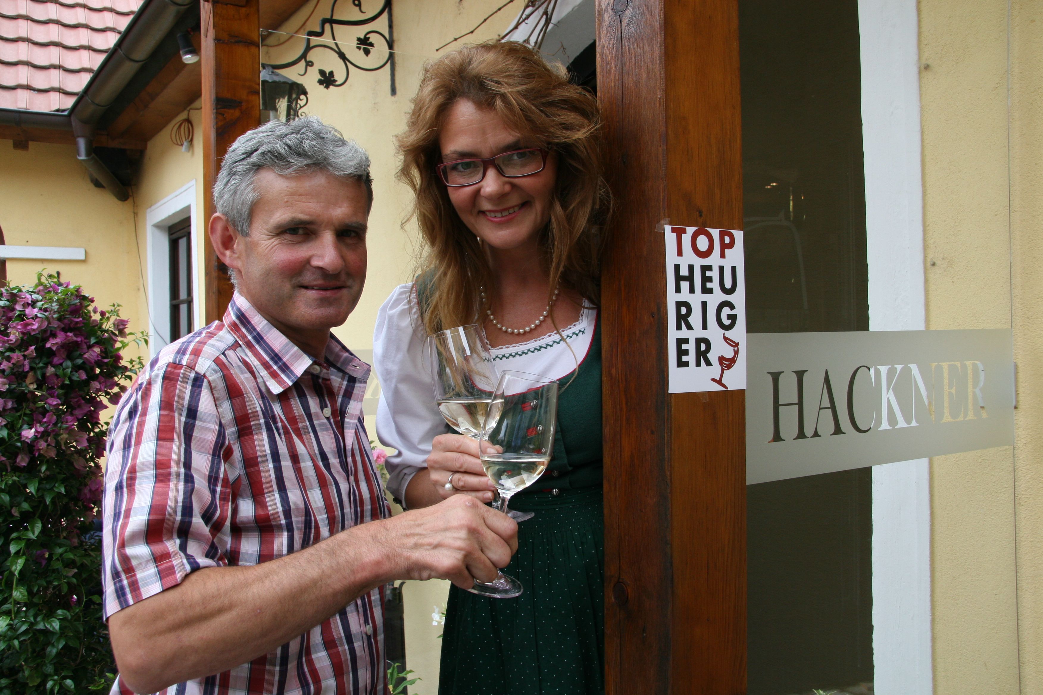 Ein Mann und eine Frau stehen vor einem Gebäude mit einem Schild 'Top Heuriger' und halten Weingläser.