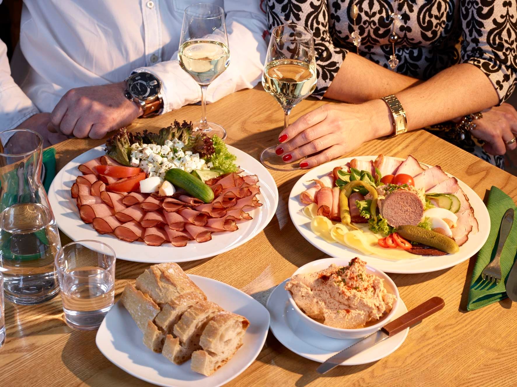 Zwei Personen sitzen an einem Tisch mit Wurst- und Käseplatten, Brot und Wein.