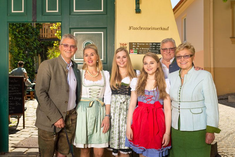 Eine Familie in traditioneller Tracht vor einem Gebäude mit der Aufschrift 'Flaschenweinverkauf'.