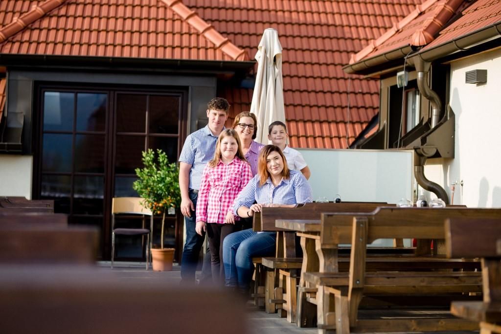 Eine Familie posiert auf einer Terrasse mit Holztischen und roten Dachziegeln im Hintergrund.