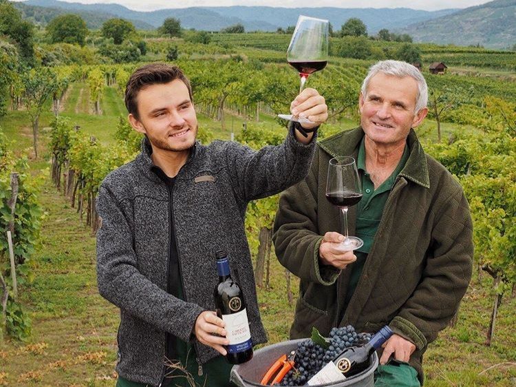 Zwei Männer in einem Weinberg halten Weingläser mit Rotwein und lächeln. Einer hält eine Weinflasche, im Hintergrund sind Reben und Hügel zu sehen.