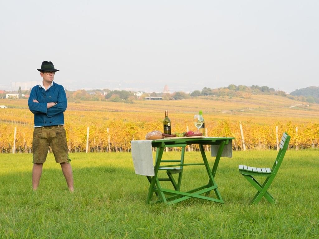 Mann in traditioneller Kleidung steht auf einer Wiese vor einem Tisch mit Weinflaschen und Brot, im Hintergrund Weinberge.