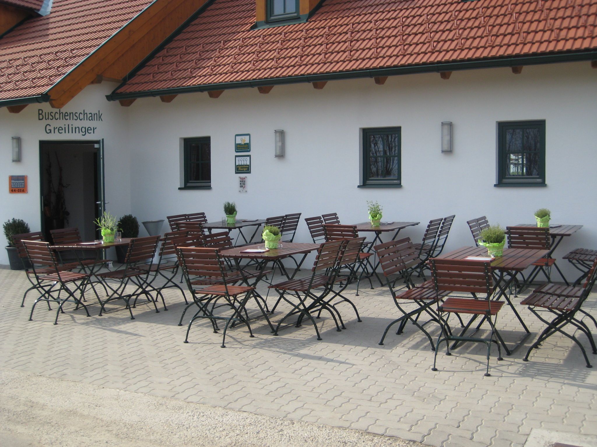 Außenbereich eines Buschenschanks mit leeren Holztischen und -stühlen vor einem Gebäude mit rotem Ziegeldach.