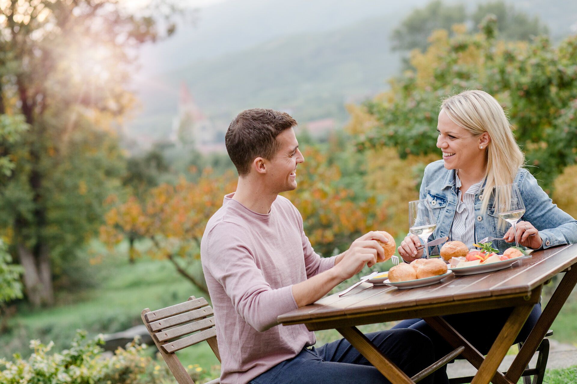 An einem malerischen Tisch im Freien genießen zwei Gäste köstliche Speisen und erfrischende Getränke. Umgeben von der idyllischen Landschaft der Wachau, strahlt die Atmosphäre eine entspannte Sommerfreude aus, während die sanften Hügel im Hintergrund die perfekte Kulisse bieten.