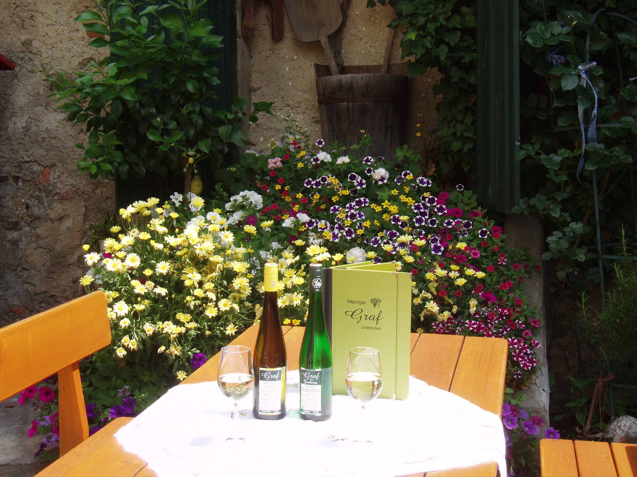 Ein Tisch im Freien mit Weinflaschen, Gläsern und einer Speisekarte, umgeben von bunten Blumen.