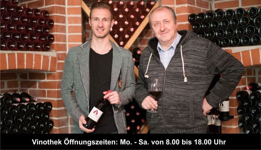 Zwei Männer in einer Weinkellerei, umgeben von Weinflaschen, einer hält ein Glas Wein.