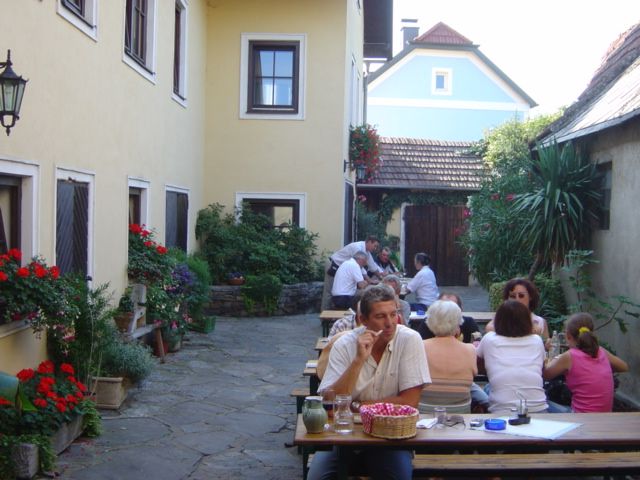 Menschen sitzen an Holztischen in einem Innenhof mit gelben Gebäuden und Pflanzen.