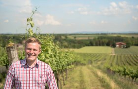 Ein l&auml;chelnder Mann steht in einem Weinberg mit weitem Blick &uuml;ber die Landschaft.