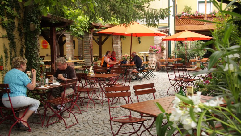 Ein gemütlicher Biergarten mit Holztischen, roten Stühlen und orangefarbenen Sonnenschirmen. Menschen sitzen und essen im Freien.