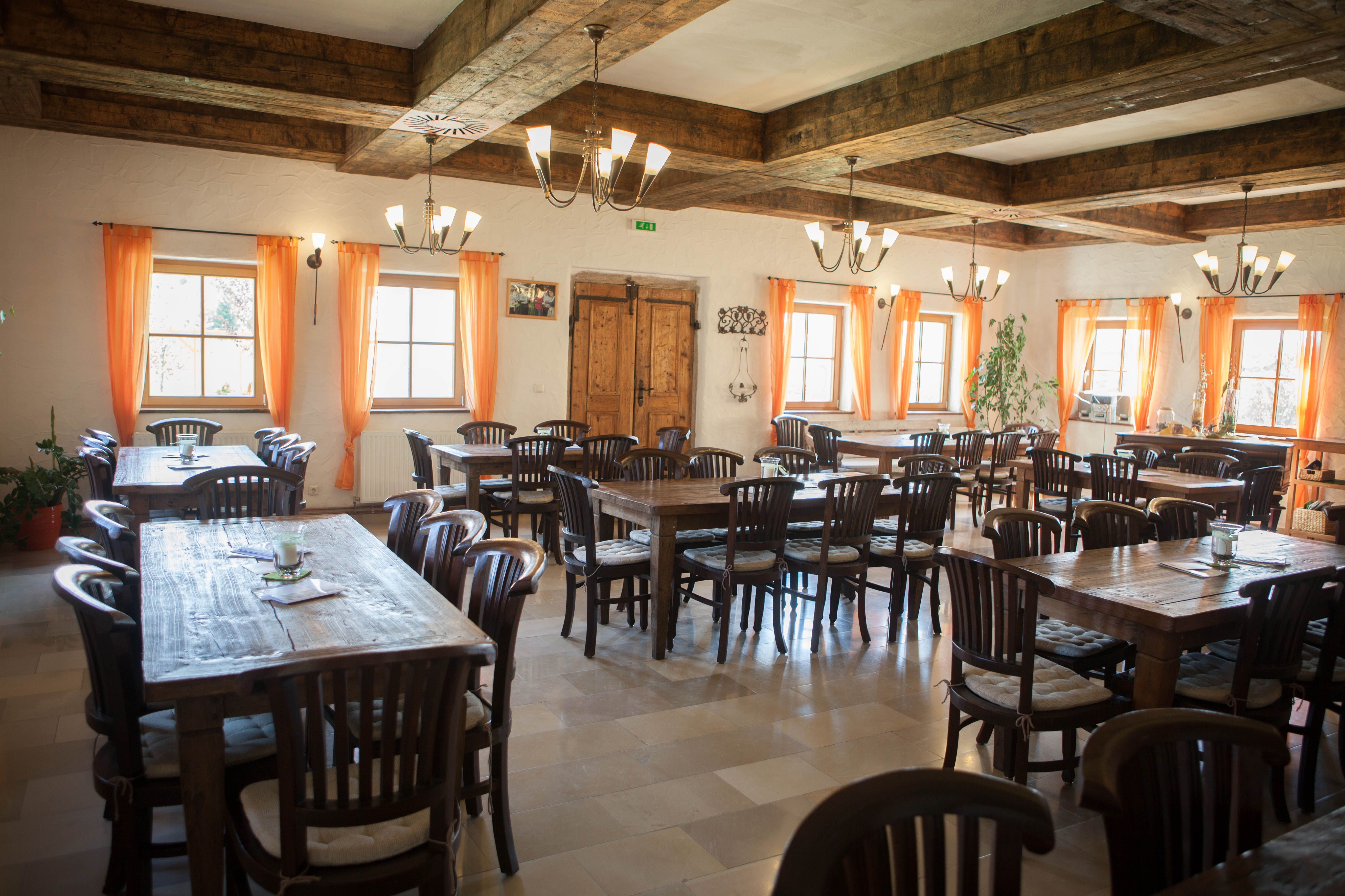 Ein rustikaler Gastraum mit Holztischen und -stühlen, orange Vorhängen und Holzbalkendecke.