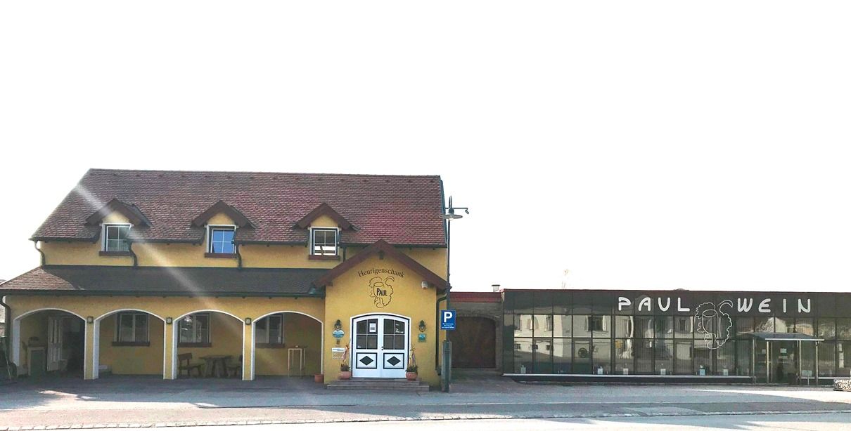 Außenansicht eines gelben Heurigenlokals mit dem Namen 'Paul' und einem modernen Anbau mit der Aufschrift 'Paul Wein'.