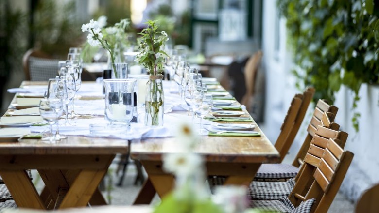 Langer Holztisch im Freien, gedeckt mit Gl&auml;sern, Besteck und Blumen.