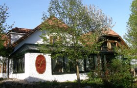 Au&szlig;enansicht eines traditionellen Geb&auml;udes mit roten Dachziegeln und einem gro&szlig;en L-Logo an der Wand, umgeben von B&auml;umen und Pflanzen.