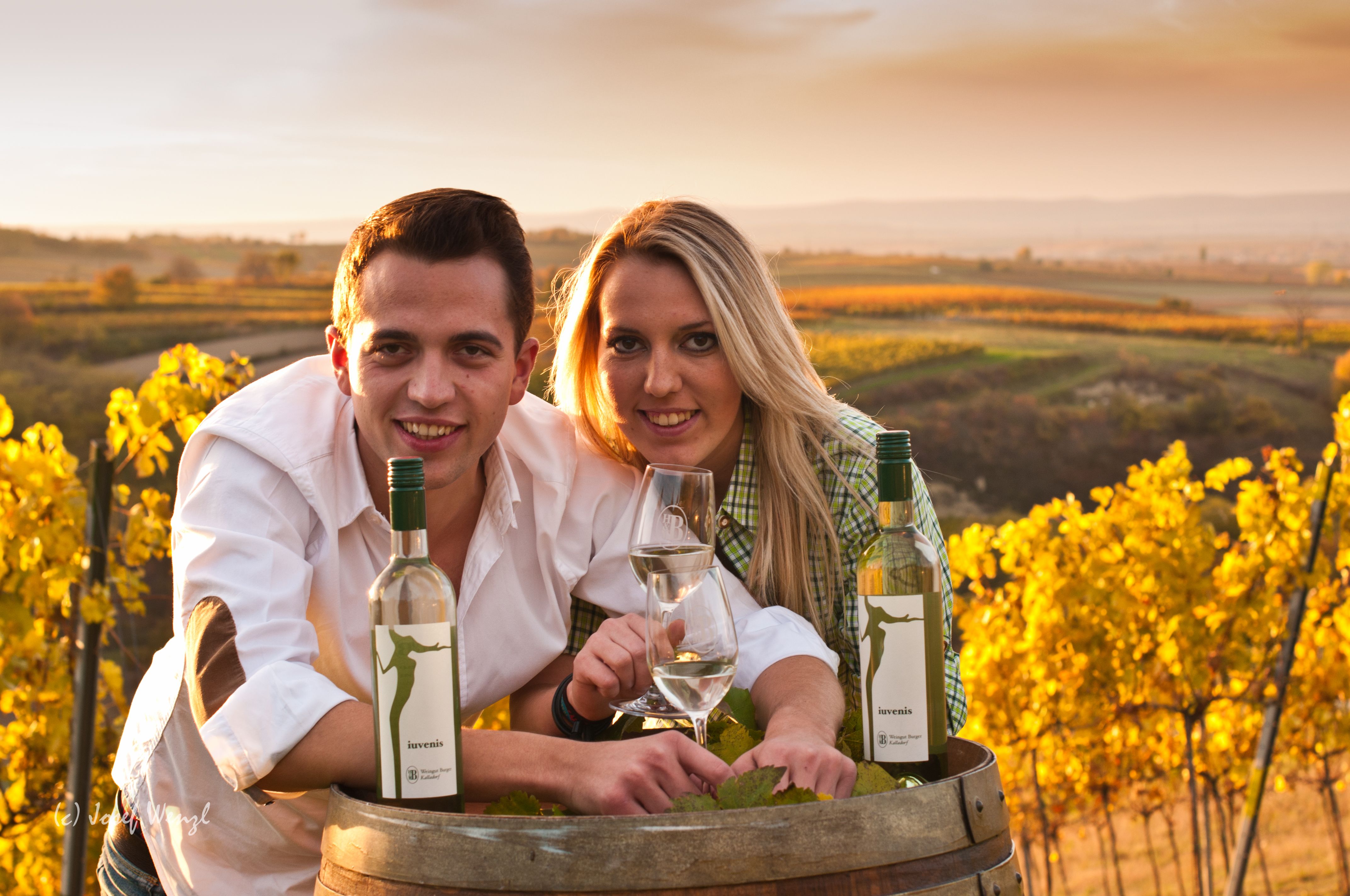 Zwei Personen lehnen sich über ein Weinfass in einem Weinberg bei Sonnenuntergang.