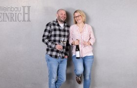 Zwei Personen stehen l&auml;chelnd vor einer grauen Wand mit Weingl&auml;sern in der Hand. Links steht ein Mann, rechts eine Frau. Oben links steht 'Weinbau Heinrich'.