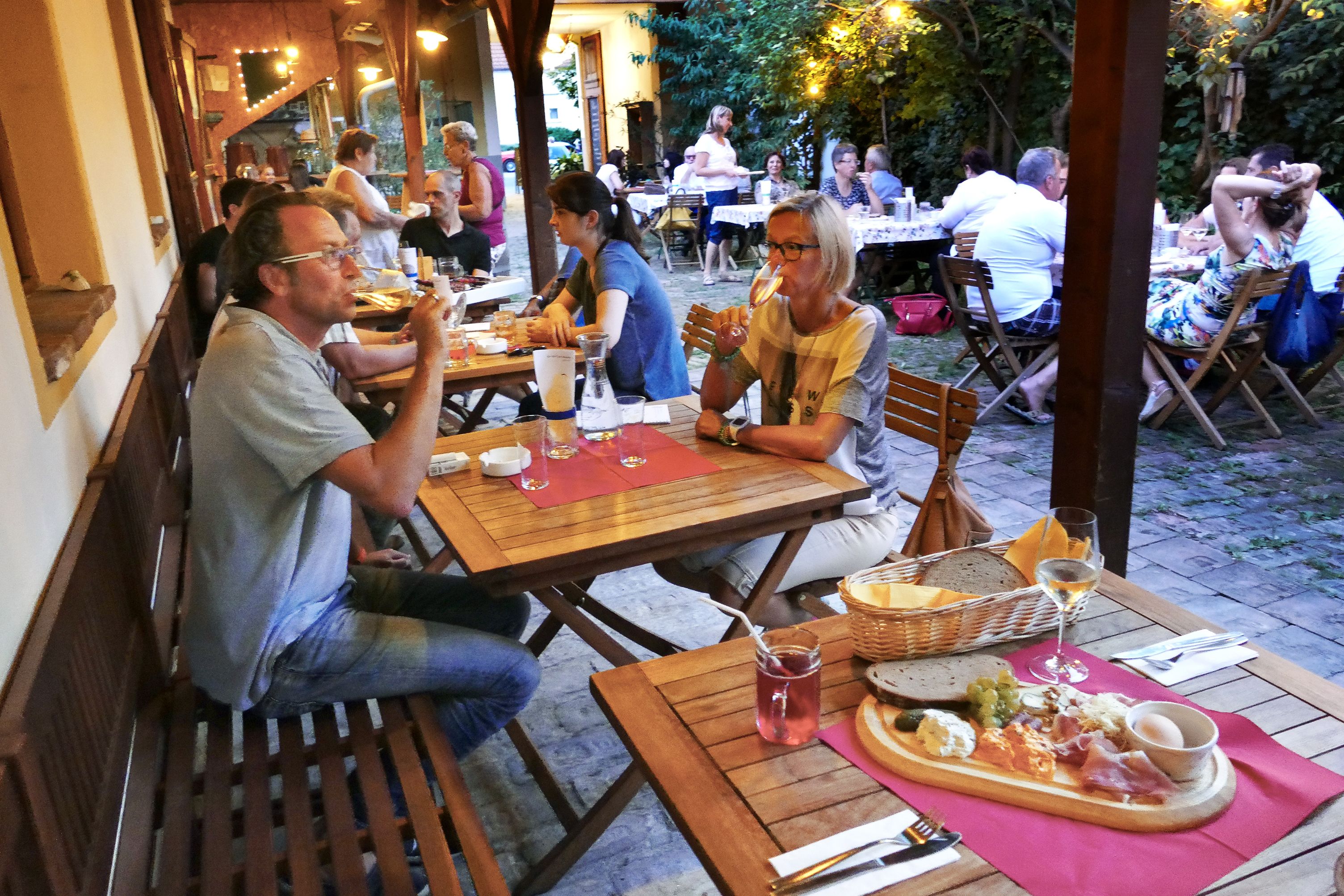 Menschen sitzen an Holztischen in einem gemütlichen Außenbereich und genießen Essen und Getränke.