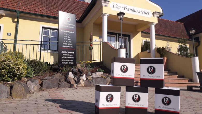 Außenansicht eines gelben Gebäudes mit der Aufschrift 'Der-Baumgartner'. Vor dem Eingang stehen Kartons mit dem Logo 'BJ'.