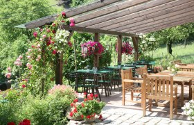 Ein gem&uuml;tlicher Au&szlig;enbereich eines Heurigen mit Holztischen und -st&uuml;hlen, umgeben von bl&uuml;henden Pflanzen und einem h&ouml;lzernen Pergola-Dach.