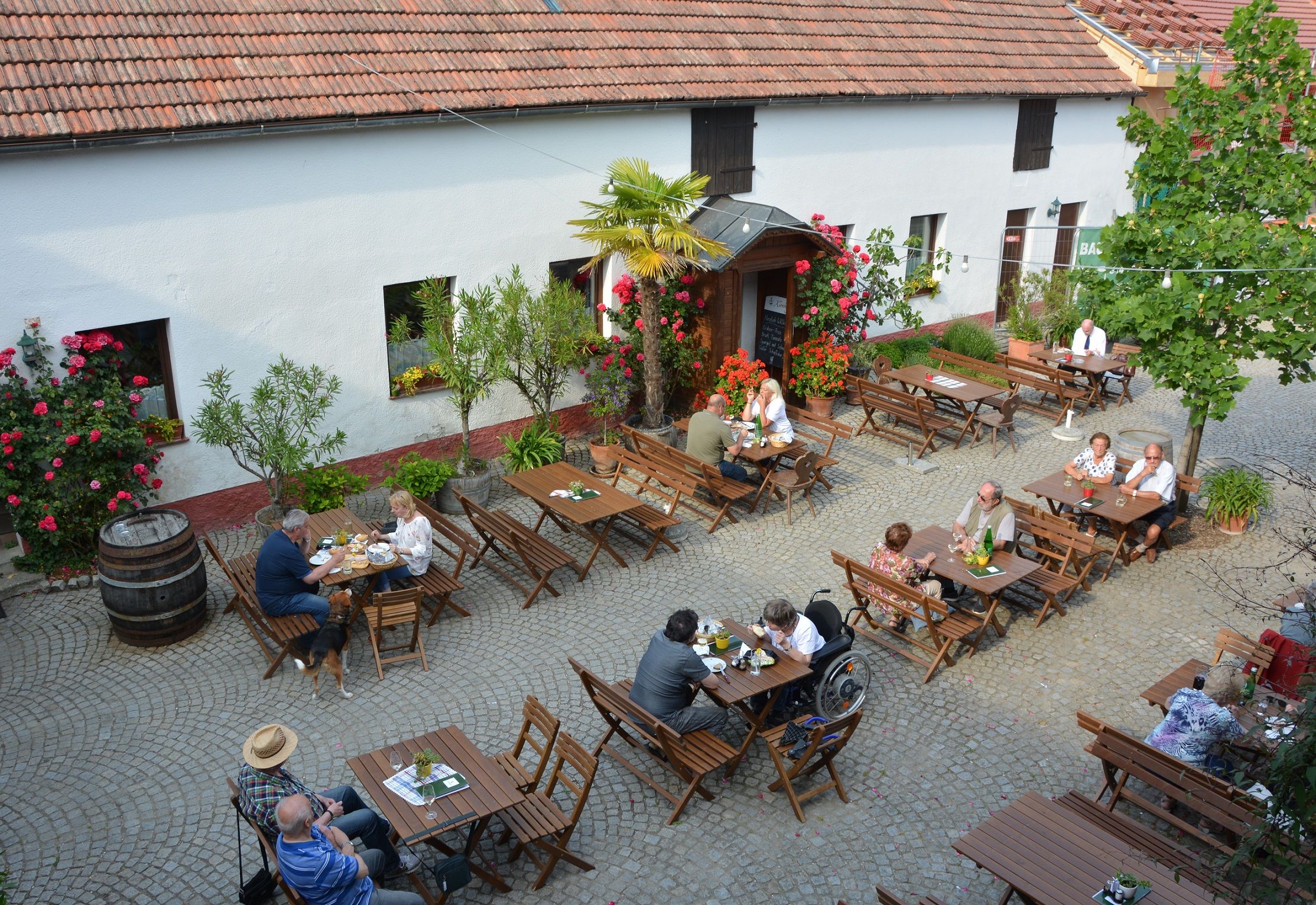 Menschen sitzen an Holztischen in einem gepflasterten Innenhof eines Heurigen, umgeben von Blumen und Bäumen.