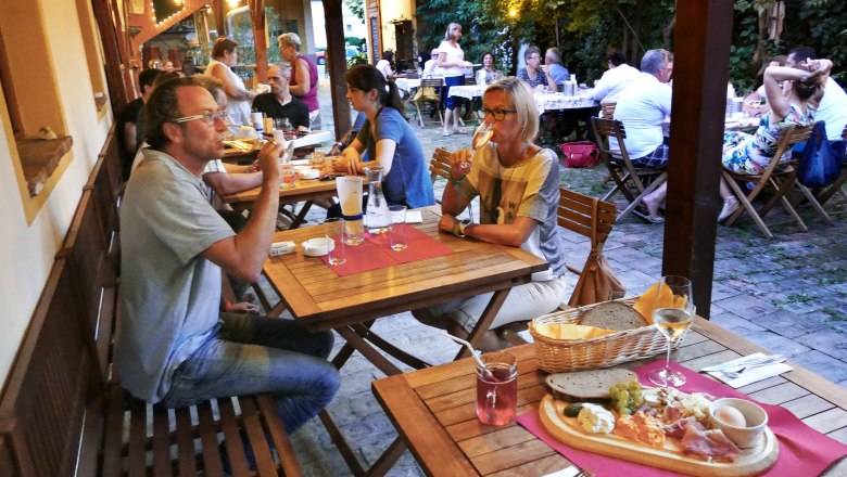 Menschen sitzen an Holztischen in einem gem&uuml;tlichen Au&szlig;enbereich und genie&szlig;en Essen und Getr&auml;nke.