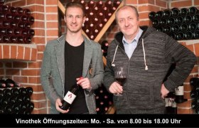 Zwei M&auml;nner in einer Weinkellerei, umgeben von Weinflaschen, einer h&auml;lt ein Glas Wein.